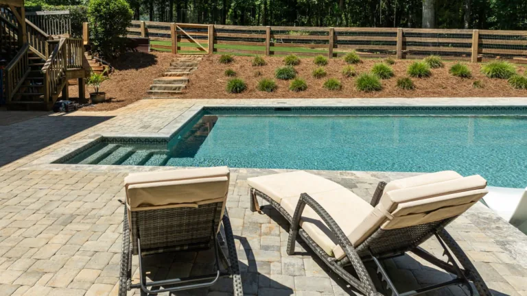 Backyard pool with lounge chairs, paver patio, and landscaped fence line under bright sun