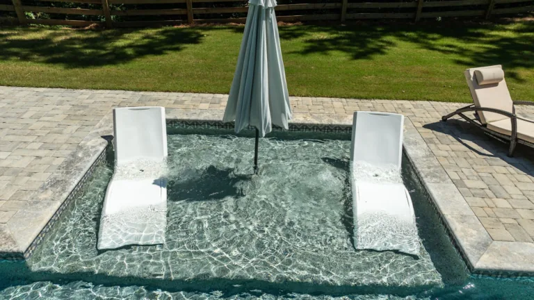 Two white lounge chairs in a small pool with a closed umbrella on a sunny patio