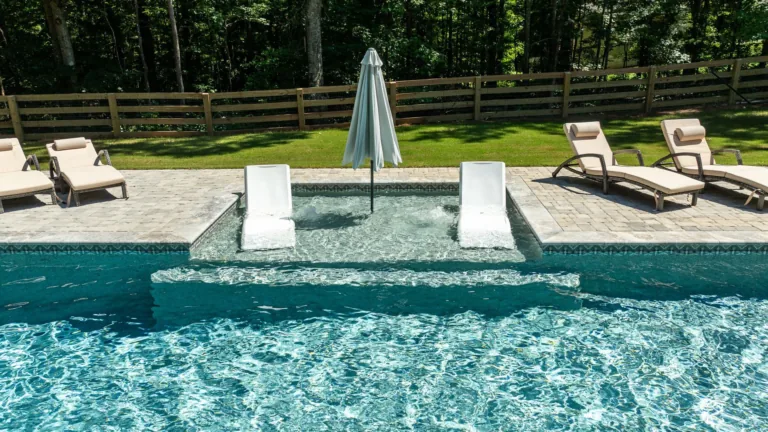 Backyard pool with two in-water loungers, umbrella, and deck chairs by a wooden fence