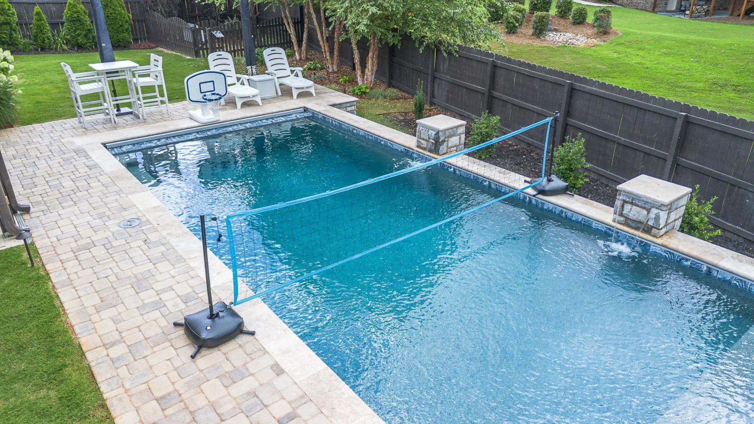 Backyard pool with a volleyball net, seating, and paved patio.