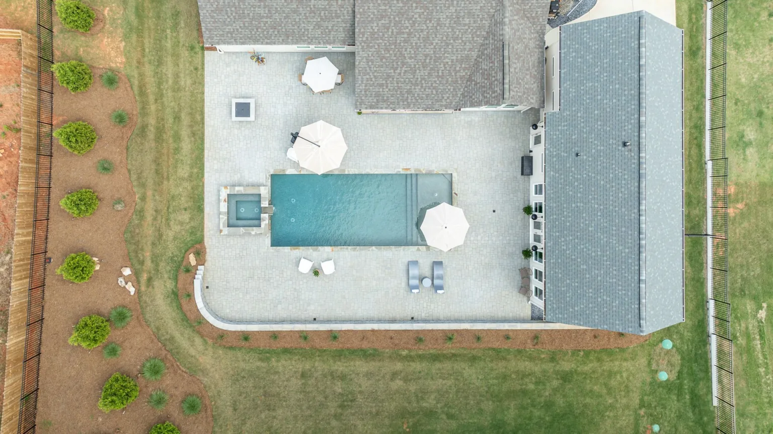 Aerial view of a backyard patio with table, umbrellas, a house, and surrounding grass and garden rows.