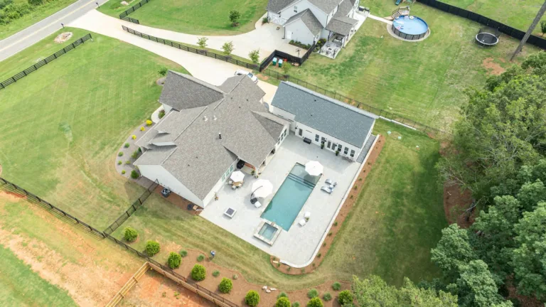 Aerial view of a suburban house with a backyard pool, patio, lawn, and surrounding trees.
