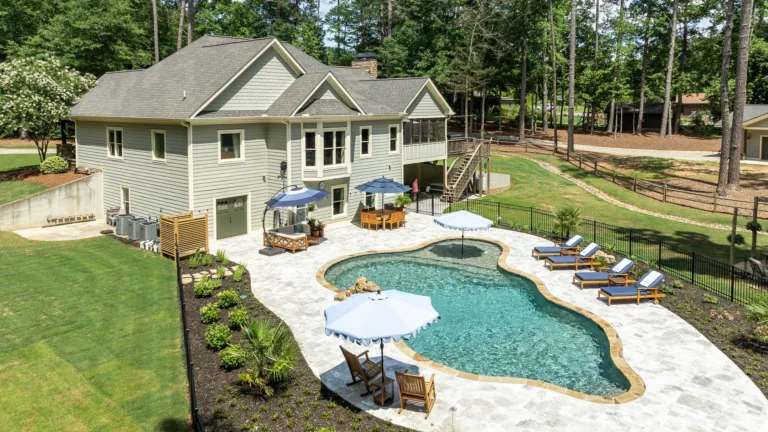Backyard with kidney-shaped pool, white stone patio, loungers, and a beige house surrounded by trees
