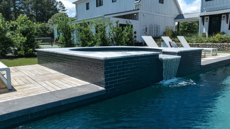 Modern backyard pool with dark tiled edge, lounge chairs, and a white house in the background