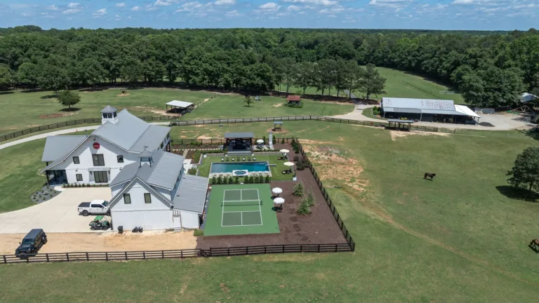 Aerial view of a large white house, lawn, and fenced tennis courts beside a tree-lined field