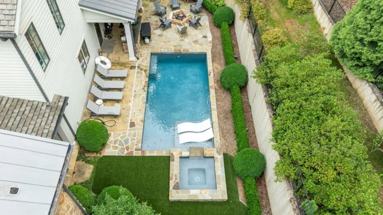 Aerial view of a backyard pool with lounge chairs, hot tub, patio seating, and green landscaping