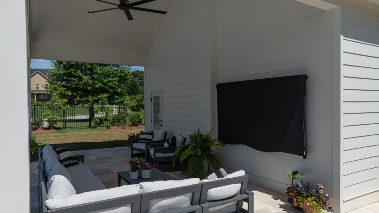 Covered patio with gray outdoor sofa, black wall-mounted TV, and potted plants beside a white house wall
