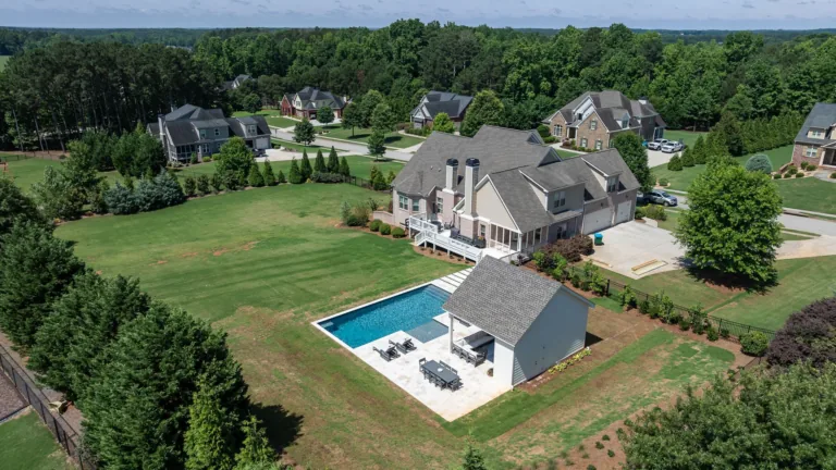 Aerial view of a suburban home with a backyard pool, patio, and surrounding green lawns and trees