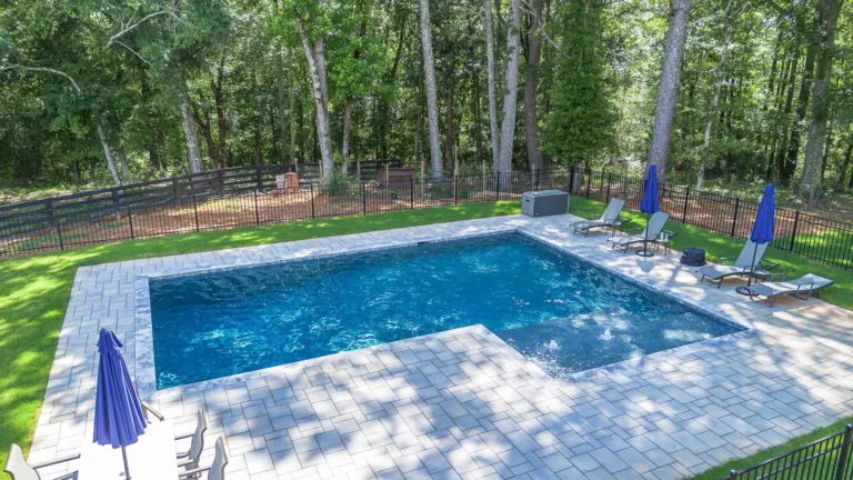 Pool with blue water, grey patio, lounge chairs, and blue umbrellas, surrounded by trees and a black fence.
