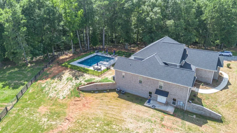 Aerial view of a brick house with a pool, surrounded by trees and grassy land.