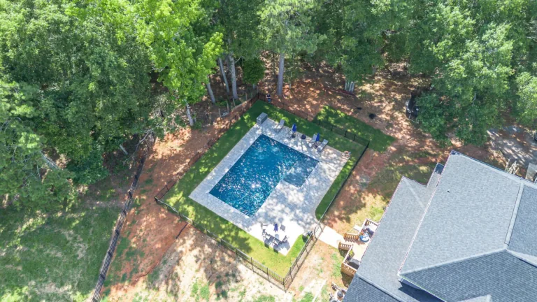 Aerial view of a blue rectangular pool surrounded by grass and trees, next to a house with a gray roof.