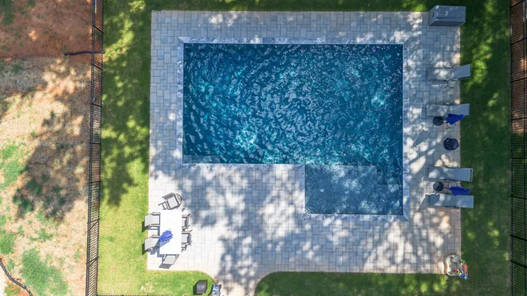 Overhead view of a rectangular swimming pool with blue water surrounded by a paved patio and green grass.